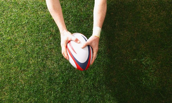 Two hands holding a rugby ball over grass.